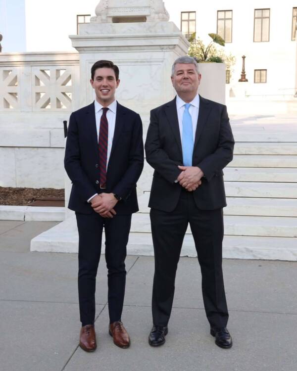 Two men, one young with a red striped tie and one older with a blue tie, smile while standing in front of a white building with steps.
