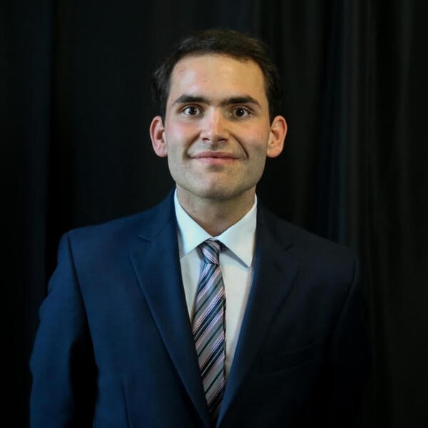 Young man in a blue suit, white shirt, and a grey, pink, and black striped tie, smiling with wide eyes.