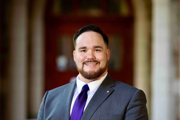 Man with dark hair and a beard, wearing a grey suit, purple tie, and Notre Dame lapel pin, smiles. Blurry red interior.