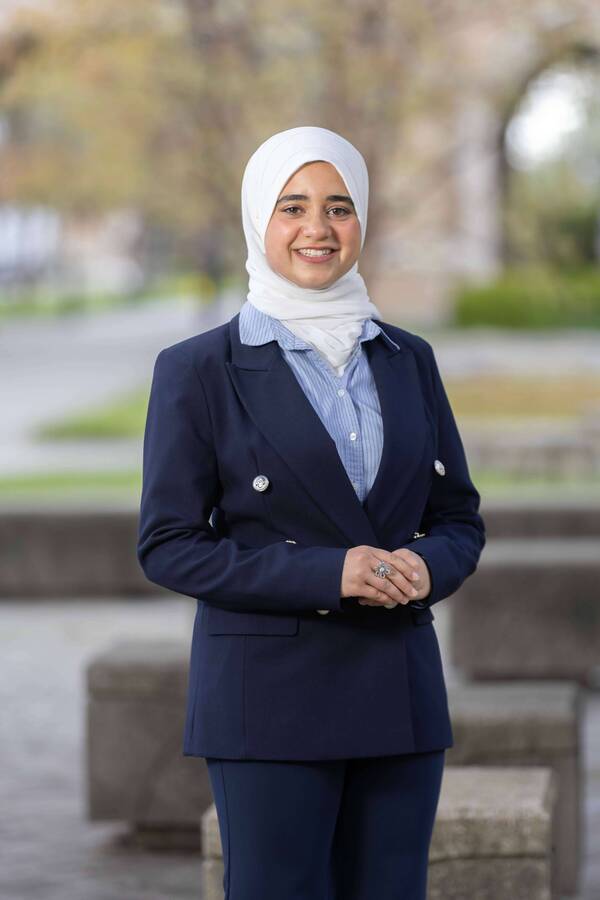 Smiling young woman in a white hijab, blue striped shirt, and navy blazer, hands clasped, outdoors.