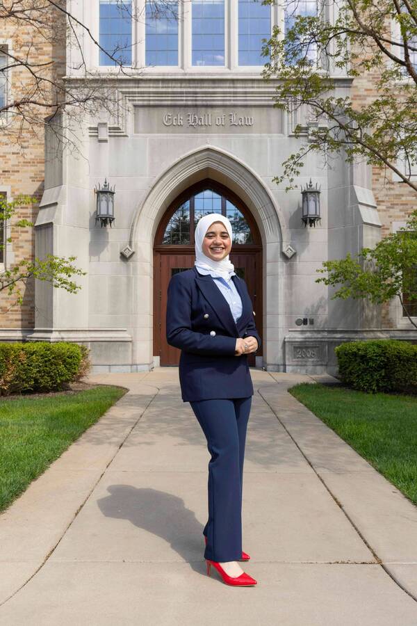 Young woman in white hijab, navy suit, and bright red heels smiles warmly in front of Eck Hall of Law.