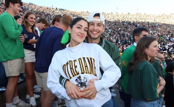 A smiling woman in a Notre Dame sweatshirt embraces a man in a green jacket and cap at a crowded stadium.