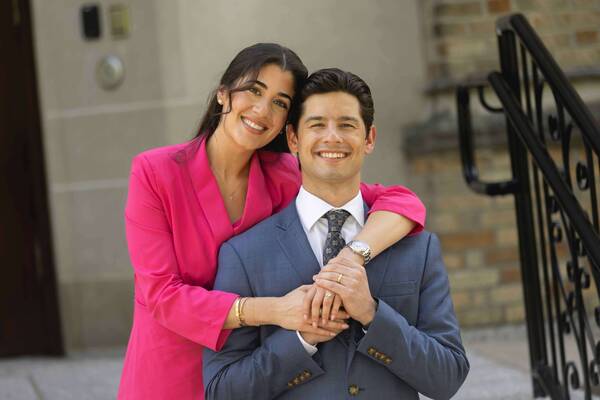 A smiling woman in a pink blazer embraces a man in a blue suit with an ND tie, hands clasped.