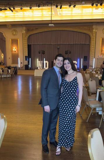 A smiling man in a dark suit and a woman in a black polka dot dress stand in an elegant ballroom with illuminated "ND" letters.