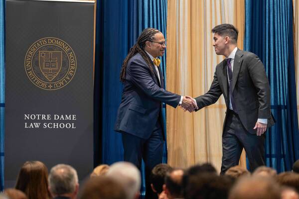 Two men shake hands and smile. One wears a navy suit and yellow bow tie; the other, a grey suit and purple tie. Notre Dame Law School.