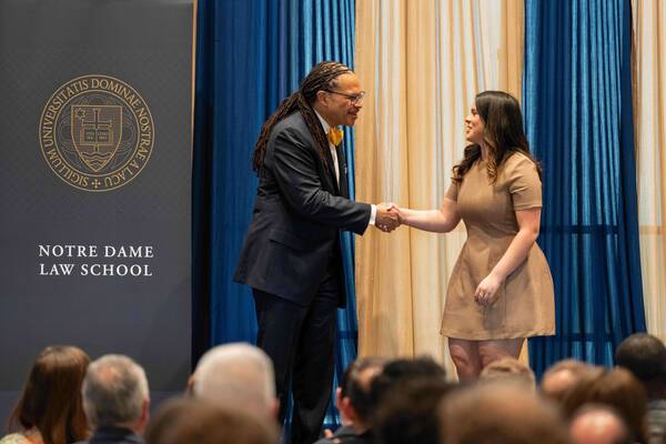 A man with dreadlocks and yellow bow tie smiles, shaking hands with a woman in a brown dress. Notre Dame Law School banner.