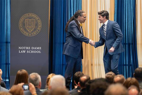 Two smiling men in dark suits shake hands. One has dreadlocks and a yellow bow tie. Notre Dame Law School banner.