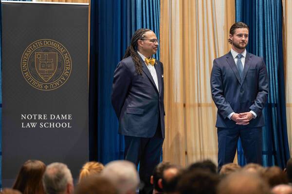 Two men in blue suits stand. Left, a man with dreadlocks and a yellow bow tie smiles left. Right, a bearded man clasps hands. Notre Dame Law School banner.