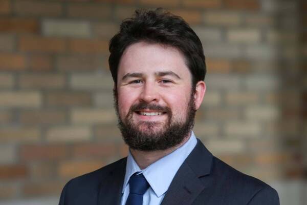 Smiling man with dark hair and full beard wears a dark suit, light blue shirt, and blue tie, against a brick background.