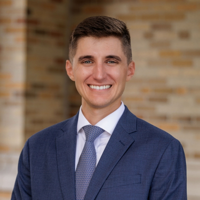 A man with short dark hair, blue eyes, and a broad smile, wearing a blue suit and patterned tie, against a light brown brick wall.