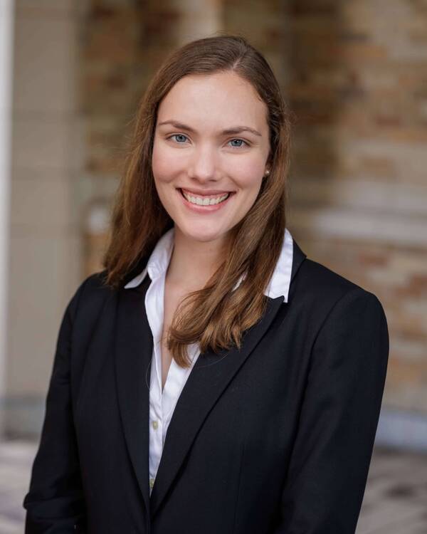 Smiling young woman with brown hair, blue eyes, wearing a black blazer and white shirt, against a blurred brick building.