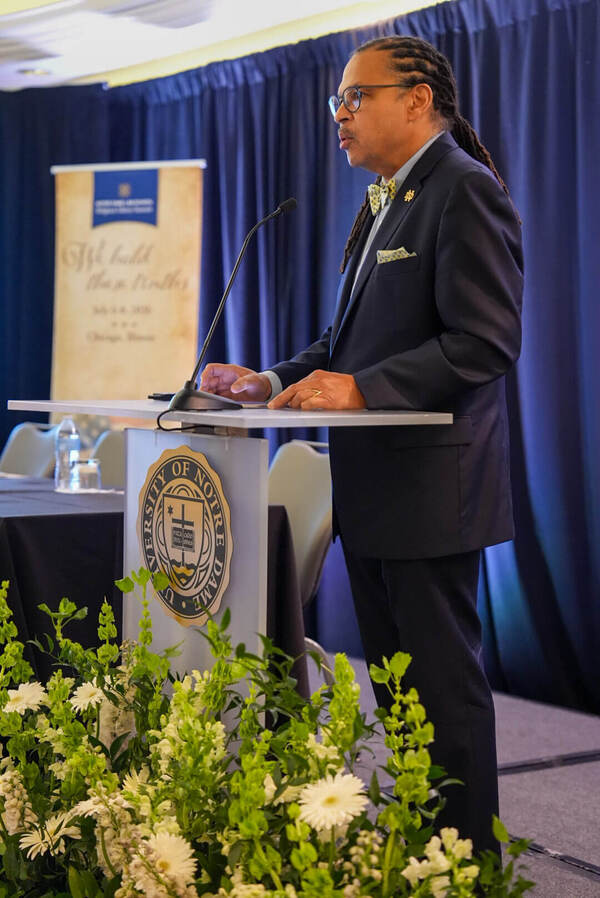 A man with glasses, navy suit, and yellow bow tie speaks at a podium with the University of Notre Dame seal.