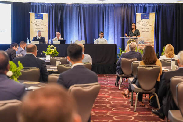 Woman in dark dress speaks at podium, gesturing. Three men sit at a panel table under a Notre Dame Law School banner.