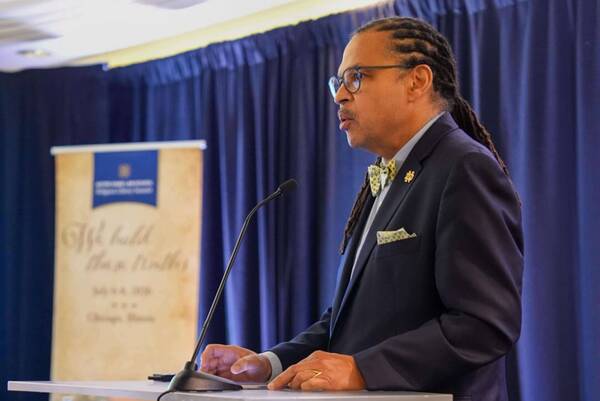 Man with dreadlocks, glasses, dark suit, yellow bow tie, and Notre Dame pin speaks at a podium with a microphone. ND banner behind him. (Dean Cole)