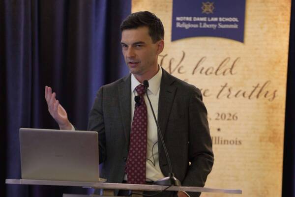 Man in a grey suit, white shirt, and red tie speaks at a podium, gesturing with his hand. Banner: Notre Dame Law School Religious Liberty Summit.