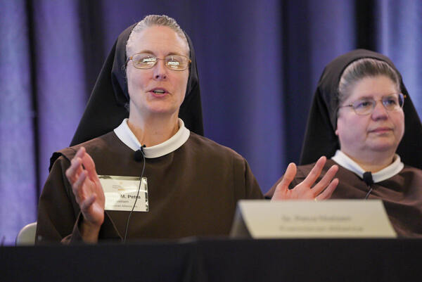 Sister M. Petra Nielsen, in brown habit and glasses, speaks into a mic, gestures.