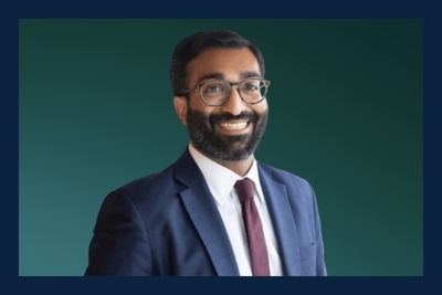 A man with a dark beard and glasses smiles broadly, wearing a navy suit, white shirt, and burgundy tie against a green background.