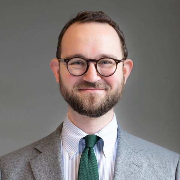 A smiling man with a beard and dark-rimmed glasses. He wears a grey blazer, light blue striped shirt, and dark green knit tie.