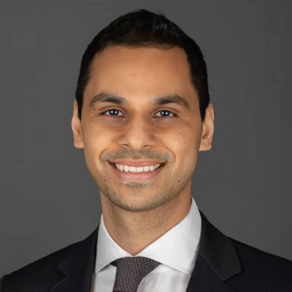 A man with dark hair, a white shirt, dark suit, and patterned tie smiles broadly against a gray background.