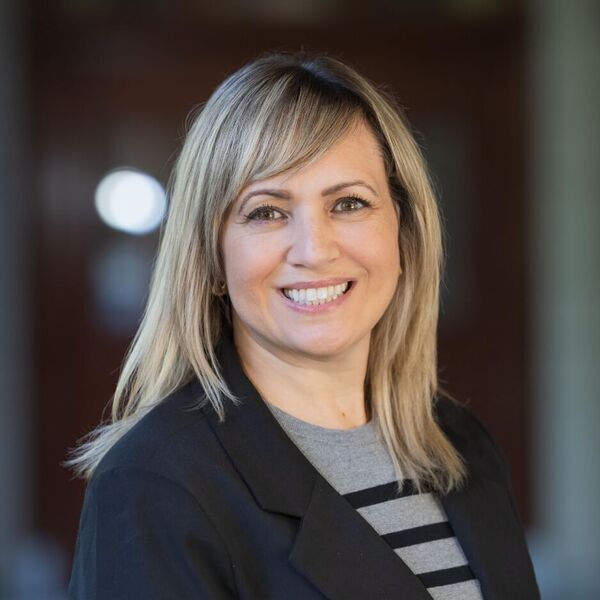 Smiling woman with light brown hair, wearing a black blazer over a grey and black striped shirt, looking friendly.
