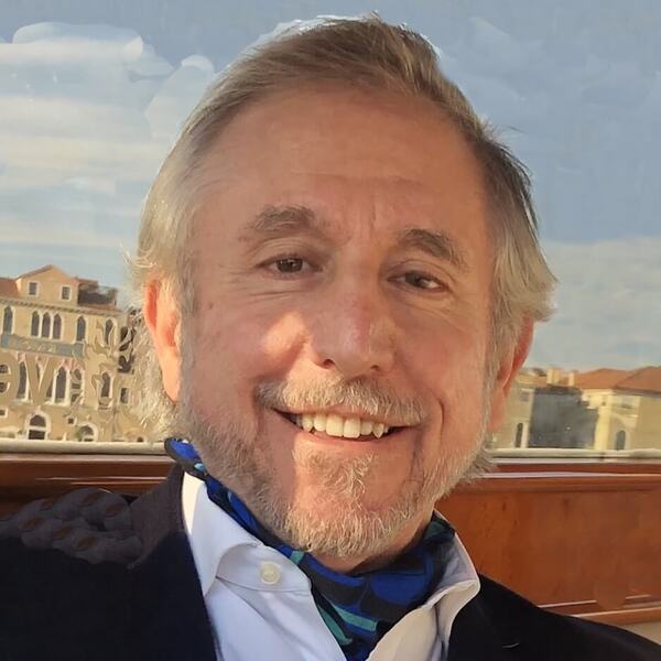 Smiling man with graying hair and beard in a blue cravat and dark jacket, against a cityscape of old buildings.