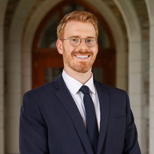 Smiling man with red hair, beard, and glasses in a navy suit against a Notre Dame stone archway.