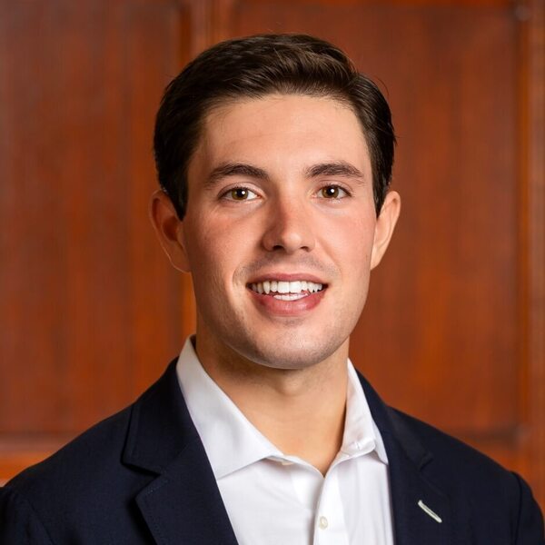 Smiling young man with brown hair and eyes in a navy suit jacket and white shirt, against a wooden background.