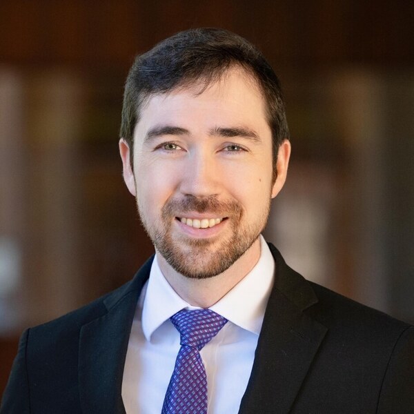 A smiling man with dark hair, a beard, and light eyes wears a black suit, white shirt, and blue patterned tie.