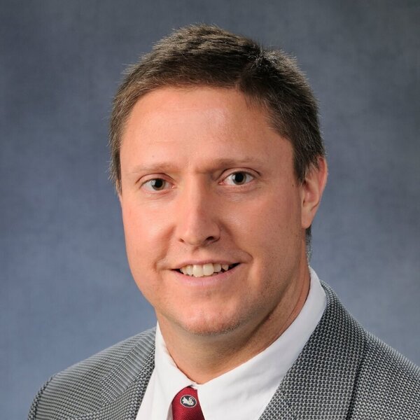 A man with light brown hair and a friendly smile, wearing a white shirt, red tie with a white squirrel, and grey blazer.