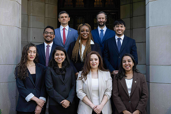 Nine smiling professionals, diverse men and women in business suits, stand in rows before a stone building entrance.