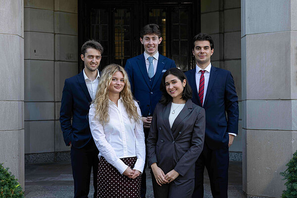 Five smiling young adults: three men in suits, two women in business attire (gray suit, polka dot skirt). Standing by a stone building.