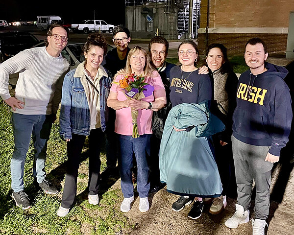Eight smiling people, including two in Notre Dame apparel, gather outdoors at night. A woman in pink holds a colorful bouquet.