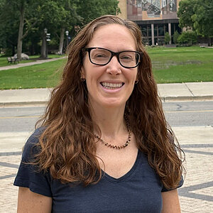 Woman with long brown hair and glasses smiles, wearing a dark blue t-shirt and necklace. (Sarah Donovan)