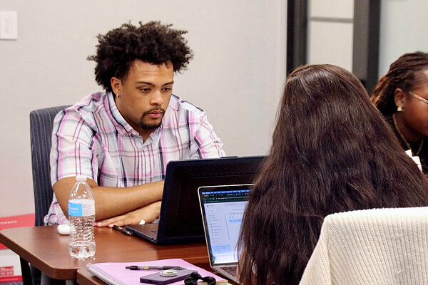 Man in pink plaid shirt focuses on laptop screen with bottle of water nearby.