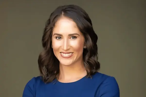 A woman with dark hair smiles, wearing a royal blue dress and subtle earrings.